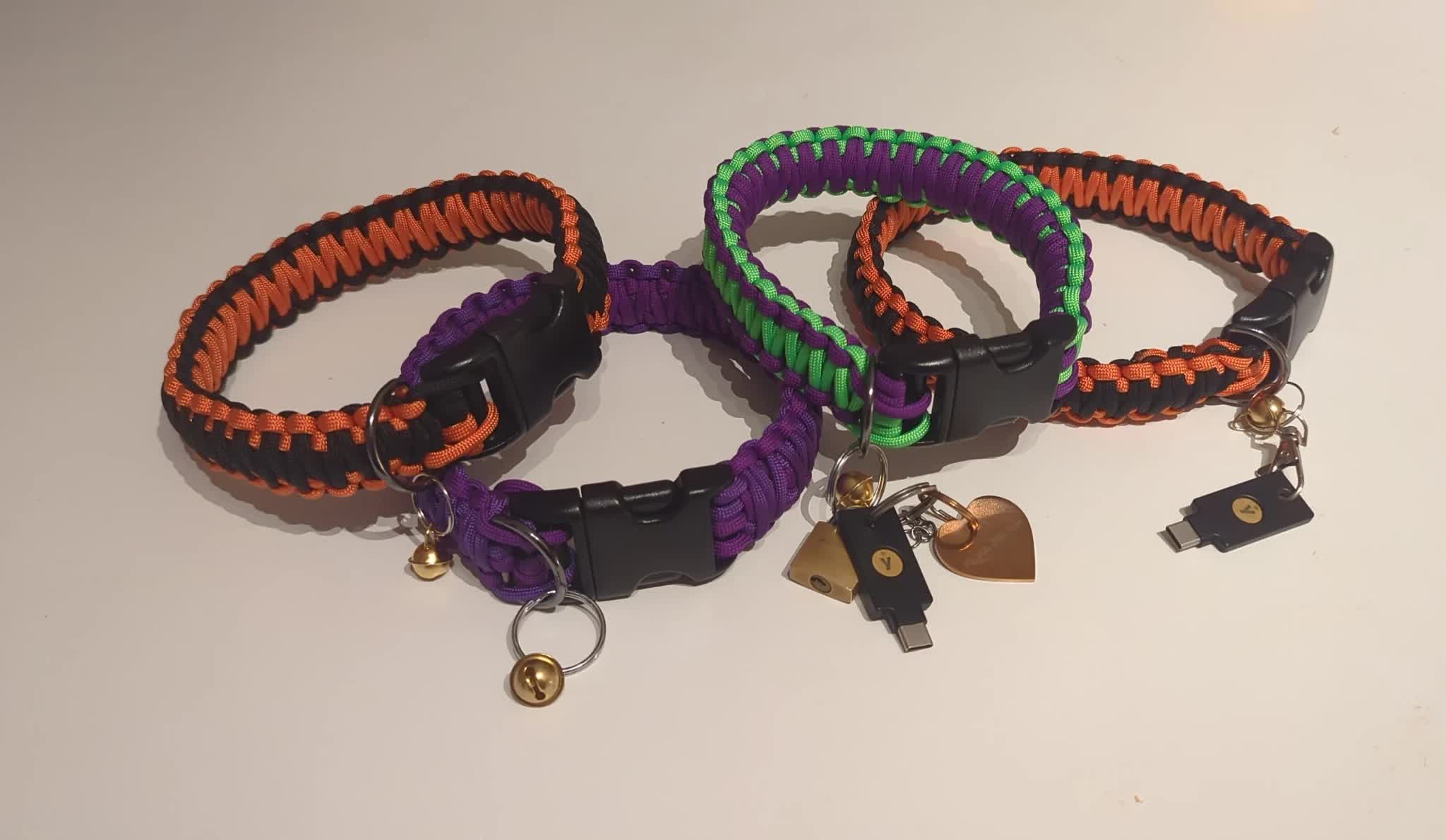 photo of 4 collars stacked on top of each other. they're made out of paracord and are colorful. several accessories are atteched to them, like bells and yubikeys.